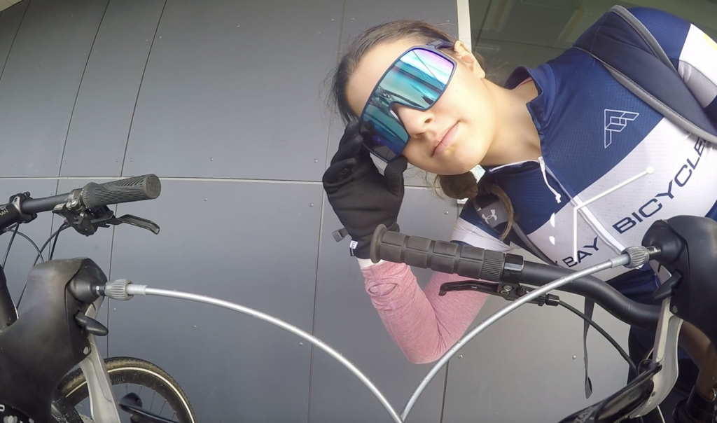 Brooke looks into her GoPro that is mounted on the handlebars of her bike. Brooke is wearing an Oak Bay Bikes Jersey over a pink long-sleeve shirt. She is wearing black gloves and blue sunglasses.